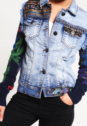 Veste en jean bleu délavé avec des motifs floraux et géométriques colorés brodés aux épaules, aux manches et aux poches, portée sur un top blanc.