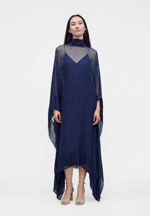 Femme portant une longue robe bleu marine transparente à col montant et manches drapées, associée à des sandales à talons hauts beiges.