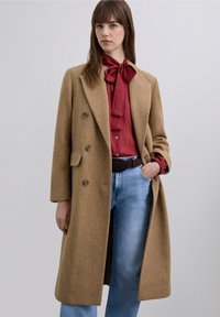 Double-breasted brown wool coat with large buttons, worn over a red blouse and blue jeans, featuring side pockets and a relaxed fit.
