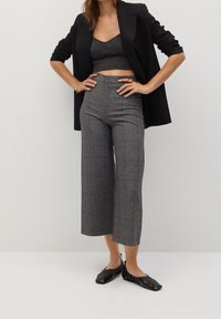 Un pantalon cropped à carreaux gris associé à un top gris assorti sous un blazer noir. Les chaussures sont des ballerines noires avec des détails froncés.
