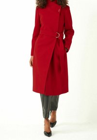 Manteau rouge avec une taille ceinturée, présentant un col asymétrique et une fente sur le côté. Texture lisse et design jusqu'aux genoux. Porté par-dessus un pantalon sombre.