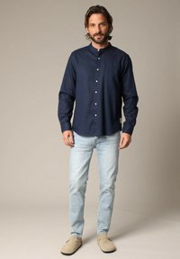 Marineblauwe button-up shirt met een mandarin-kraag, gemaakt van zachte stof. Gemaakt met lichtblauwe jeans en beige instappers, staande tegen een neutrale achtergrond.