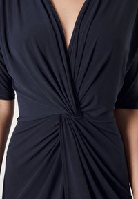 Mörkblå klänning med djup V-ringning, korta ärmar och intrikata veck vid midjan. Slätt, elastiskt tyg ger en figurnära silhuett.