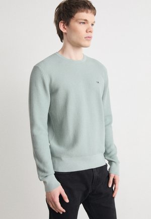Maglione a coste verde chiaro con scollatura a girocollo, polsini e orlo a costine. Piccolo dettaglio logo nero sul petto. Abbinato a pantaloni neri.