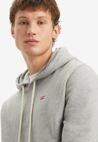 Sudadera gris hecha de una tela suave que presenta un bolsillo canguro frontal, dos cordones de color crema y un logo rojo de Levi's en el pecho.