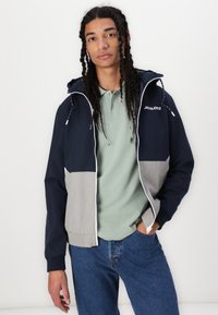 Navy- und graufarbene Farbblockjacke mit Kapuze, weißem Reißverschluss und Kordeln. Getragen über einem hellgrünen Polo und blauen Jeans.