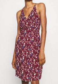 Květinové šaty v burgundské barvě, s výstřihem do V, ramínky na špagetách a nařaseným pasem. Design obsahuje vícebarevné květiny na jemném materiálu.
