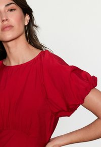 Robe rouge à manches courtes avec un col froncé et des manches volumineuses. Texture lisse avec un léger éclat et un détail de taille cintrée.