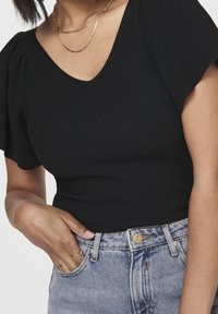 Top de punto negro con canalé que presenta un escote en V y mangas cortas con volumen, combinado con jeans de mezclilla azul claro y cadenas de oro apiladas.