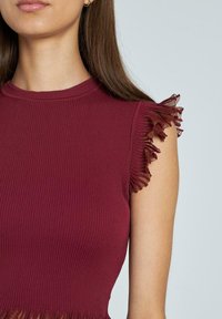 Donna che indossa un top senza maniche in maglia bordeaux con bordo in mesh arricciato sulle spalle e sull'orlo, su uno sfondo chiaro e neutro.