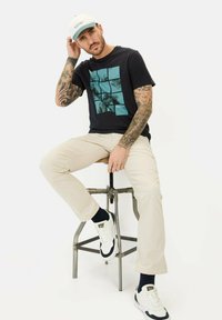 Sort T-shirt med turkise grafiske design, beige bukser og hvide sneakers. Sidder på en metalstol med en lys baggrund.