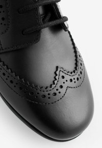 Chaussure en cuir noir avec un bout rond, des perforations décoratives et des accents en zigzag. Les lacets passent à travers de petits œillets.