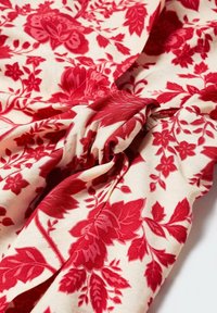 Tissu crème avec des motifs floraux et de feuilles rouges denses, légèrement froncé pour montrer la texture et les plis.