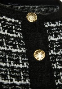 Tissu en tweed noir et blanc avec bordure tricotée noire comprenant deux boutons dorés décoratifs avec des motifs ornés.
