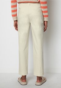 Ljust beige bomullshängsel med avslappnad passform, raka ben, två bakfickor och minimala sömdetaljer. Klädd med platta sandaler.