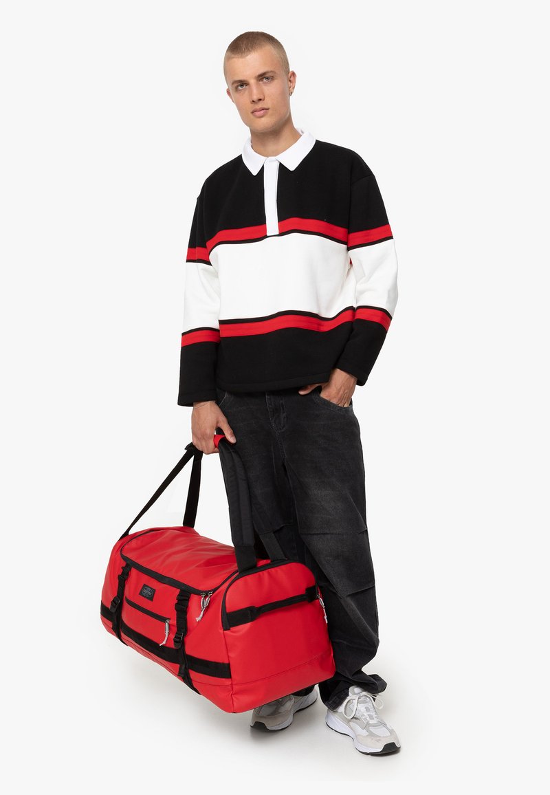 Sac de sport rouge avec des accents noirs et plusieurs poches zippées, assorti à une chemise à rayures noires et blanches à manches longues.