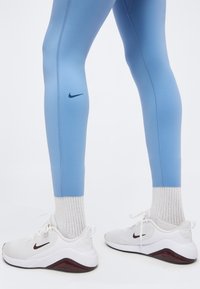 Πόδια που φορούν ανοιχτό μπλε κολάν Nike, λευκές κάλτσες και λευκά αθλητικά παπούτσια Nike με μαύρο σήμα και καφέ λεπτομέρειες, στέκονται σε λευκό φόντο.