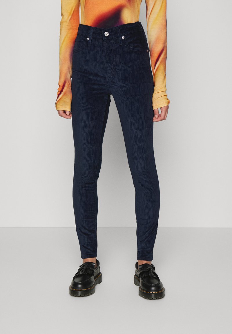 Calças de veludo cotelê azul marinho com um corte slim, cintura alta e uma textura ribbed vertical subtil. Apresenta um fecho com botão e zipper, além de bolsos laterais.