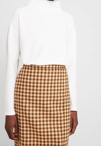 Maglione bianco testurizzato con colletto alto abbinato a una gonna a motivi houndstooth marrone e gialla. Esposto su uno sfondo grigio.