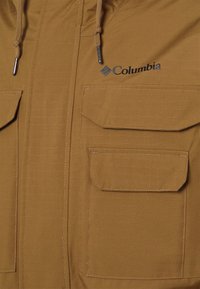 Brun, lättviktsjacka i tyg med två stora framfickor, justerbar huva och Columbia-logotyp i svart på bröstet.