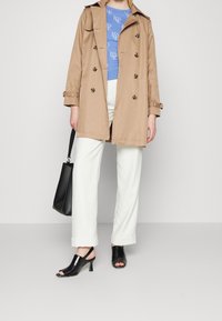 Cappotto beige con design a doppio petto, maglione a righe blu chiaro, pantaloni bianchi a gamba larga e sandali neri con tacco.