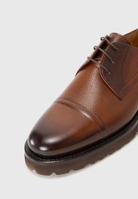 Bruine lederen gekleed schoen met een gestructureerde bovenkant, ronde neus en decoratieve stiksels. Heeft een duurzame rubberen zool en vetersluiting.