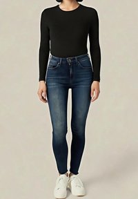 Personne portant un haut à manches longues noir, un jean skinny bleu foncé et des baskets blanches, se tenant devant un fond beige uni.