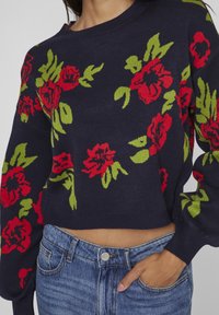 Pull marine à motif floral rouge et vert. Présente une coupe courte, un col rond et des poignets côtelés. Porté avec un jean taille haute.