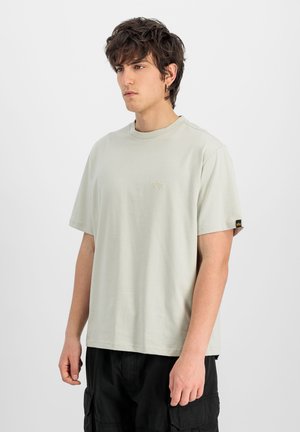 Giovane uomo con capelli castani spettinati che indossa una maglietta beige chiaro a maniche corte e pantaloni cargo neri, in piedi davanti a uno sfondo semplice.