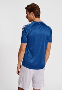 Rövid barna hajú férfi, kék rövid ujjú sportpólóval, melyen fehér chevron minták vannak a vállán, és fehér sportshortot visel, hátulról nézve.