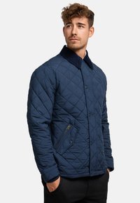 Navy quilted jacket with a corduroy collar, featuring a zippered pocket and button closure, crafted from a smooth, padded synthetic material.