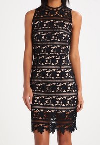 Hihaton, istuva mekko mustasta kukkakuvioiduista pitsistä, jossa on nuden värinen aluskangas, korkea kaulaosa ja aallotettu helma polvien ylle.