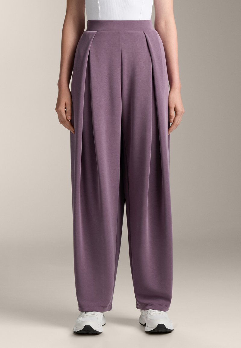 Lila weite Hosen mit Falten, aus glattem Material und mit hohem Taillenbund, kombiniert mit weißen Sneakers.