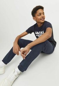 Marineblauwe trainingsbroek met geribbelde enkels en een slim fit ontwerp, gecombineerd met een zwart t-shirt met witte tekst. Witte sneakers maken de outfit af.