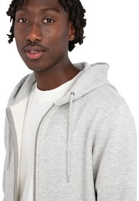 Sivá mikina na zips s jemnou textúrou, nastaviteľnou kapucňou so šnúrkou a akcentom loga, nosená cez biele tričko.