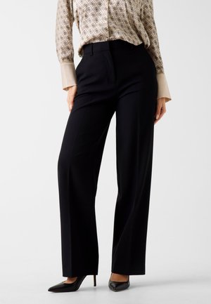 Donna che indossa pantaloni neri a vita alta e gamba larga, camicia beige fantasia con grandi polsini e scarpe nere con tacchi a punta, in piedi.