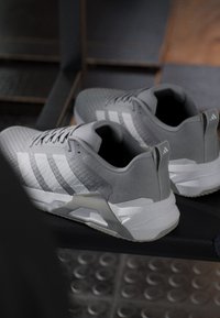 Paire de baskets grises Adidas avec des bandes blanches reposant sur un banc noir dans un vestiaire.