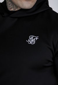 Črna kapuca izdelana iz gladkega materiala, ki ima na prsih majhen beli vezen logotip. Oblikovanje vključuje kapuco s vrvico.