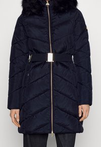 Tmavě modrý prošívaný kabát s kapucí s umělým kožíškem, prošívaným vzorem, páskem v pase a zlatým zipem. Má boční kapsy.