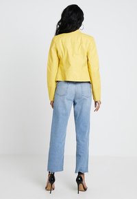 Kvinna med långt svart hår som bär en gul jacka, ljusblå jeans och svarta högklackade skor, stående vänd bort från kameran mot vit bakgrund.