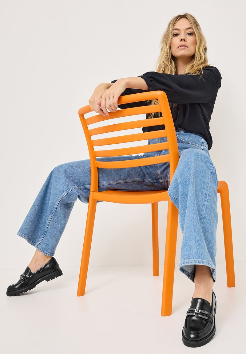 Oranje plastic stoel met horizontale latten. Model in zwarte top en blauwe flared jeans. Zwarte loafers met metalen accenten. Neutrale achtergrond.