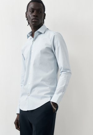 Homme portant une chemise bleu clair boutonnée et un pantalon foncé, debout avec une main dans la poche, sur fond blanc uni.