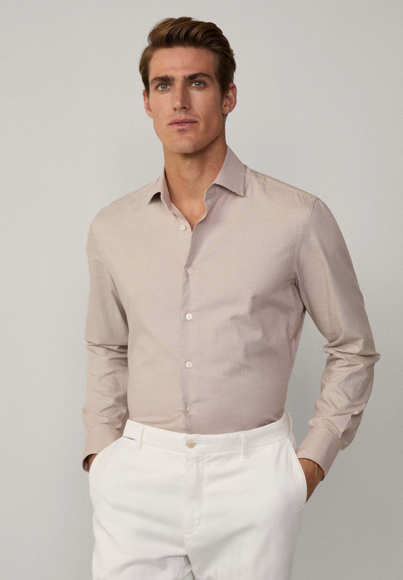 Chemise à manches longues marron clair avec un motif rayé subtil, col à boutons, et boutons blancs, associée à un pantalon blanc.