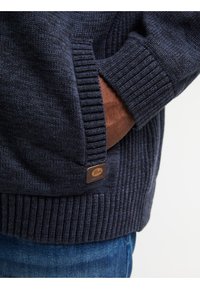 Mörkblå stickad tröja med ribbade manschetter och nederkant. Har en brun läderetikett med varumärkeslogotyp. Mjuk textur och avspänt passform.