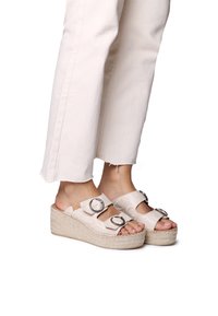 Sandali con zeppa con una tomaia beige testurizzata, due fibbie regolabili e un tacco di juta intrecciata. Indossati con pantaloni bianchi a vita corta.