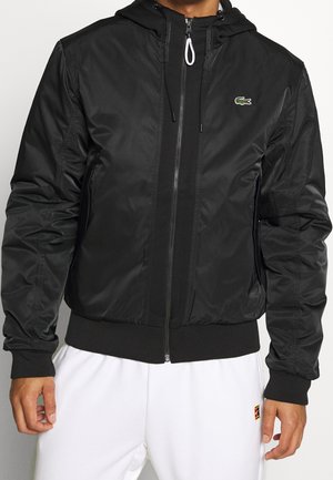 Mężczyzna w czarnej kurtce z kapturem zapinanej na zamek, z małym zielonym logo krokodyla na piersi, w białych spodniach.