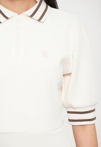 Λευκό πόλο μπλουζάκι με σχέδιο ημικαζάνας, ριμπ κολάρο με καφέ και λευκές ρίγες, και κοντά μανίκια με αντίστοιχες μανσέτες. Υποτονικό λογότυπο της μάρκας ανάγλυφο.