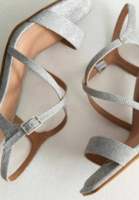 Talons paillettes argentées avec des brides croisées, partie supérieure texturée, détail de boucle et intérieur beige. Semelles plates et design à bout ouvert.