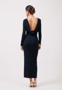 Femme vêtue d'une robe noire à manches longues, dos nu avec un dos drapé bas, portant un collier chaîne en or, des bracelets, et des sandales blanches à talons, debout de dos.