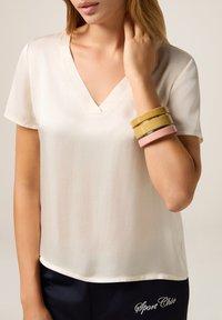 Blusa blanca de manga corta y escote en V con una textura suave. La modelo lleva pulseras coloridas: amarilla, verde, rosa y plateada. Pantalones azul marino con bordados.